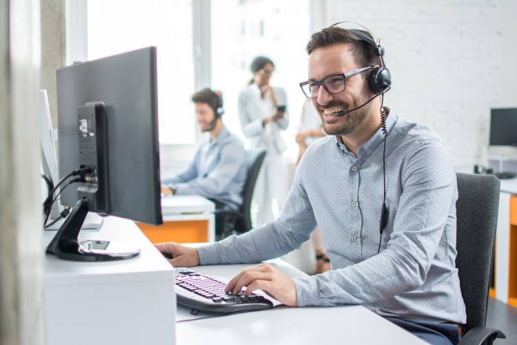 Man sitting at computer talking on a headset and smiling with co-workers in the background